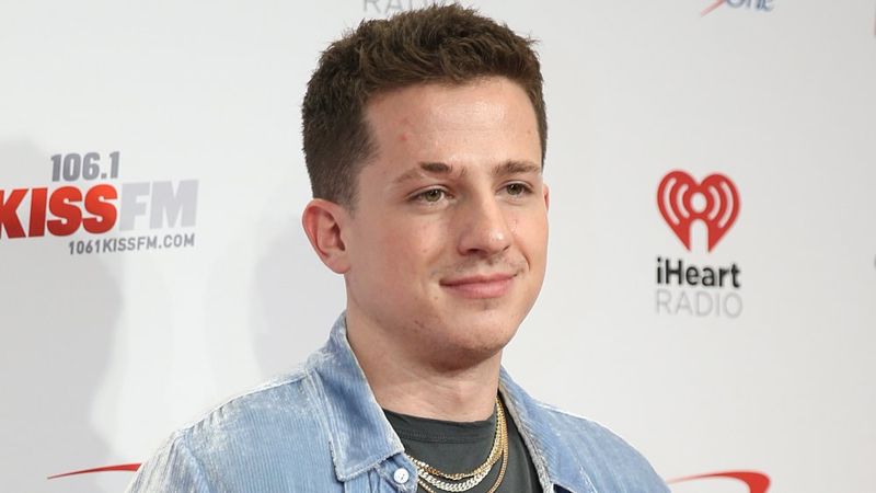 Charlie Puth em um botão de veludo cotelê azul claro, camiseta preta e correntes, posando com uma expressão neutra em um evento da iHeartRadio